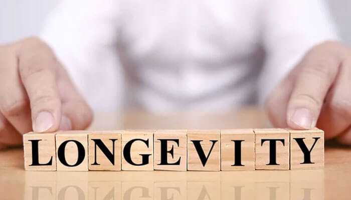 The image shows wooden blocks spelling out the word longevity symbolizing mental and physical wellness, with a persons hands arranging the blocks on a reflective surface