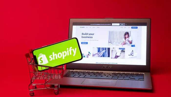 Shopify dropshipping store logo on the phone in a shopping cart, with a laptop in the background showing shopify's website.