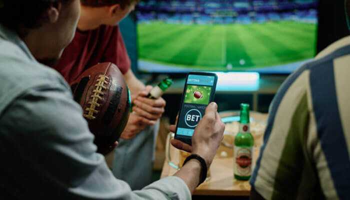 Group of friends watching a football match on tv, one using a smartphone with a betting app-foregrounding betwinner partners.