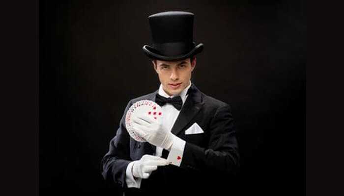 A man dressed in formal poker style, wearing a top hat, tuxedo, and white gloves, demonstrating card tricks with a deck of playing cards.
