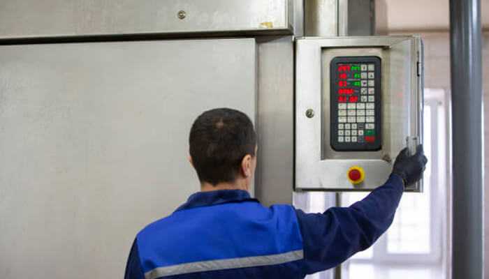 Worker adjusting a control system with load cells in a factory setting, interacting with a machine panel for operations monitoring.