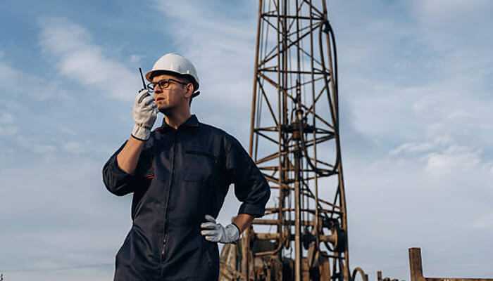 Technician in safety helmet using poclink walkie talkie near an industrial drilling rig under blue sky.