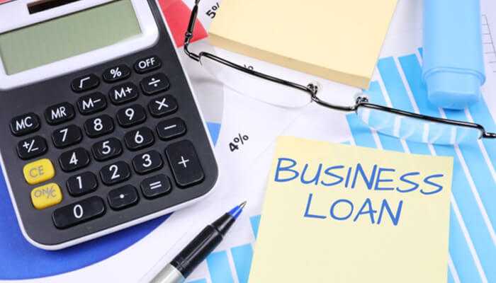 Calculator, eyeglasses, and a yellow sticky note reading “business loan” placed on financial charts, representing commercial loans and financing tools