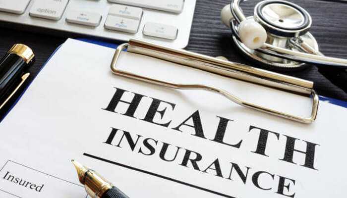 
A document on a clipboard with the bold words "HEALTH INSURANCE" printed across the center. A black pen with a gold nib and a stethoscope rest on the document, and a computer keyboard is visible in the background.