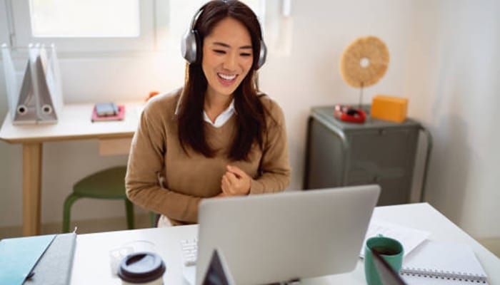 A woman smiling while working on her laptop with headphones on in a comfortable home office setting. This is a perfect example of how to make money fast as a woman through online work, such as freelancing or tutoring.