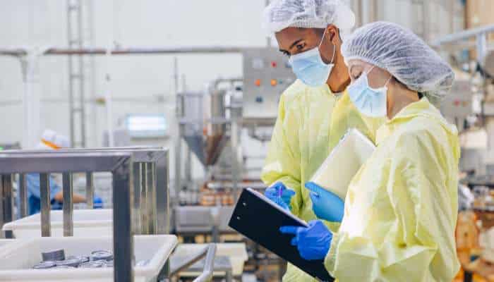 Two workers in yellow protective gowns, masks, and caps review a clipboard in a manufacturing facility.