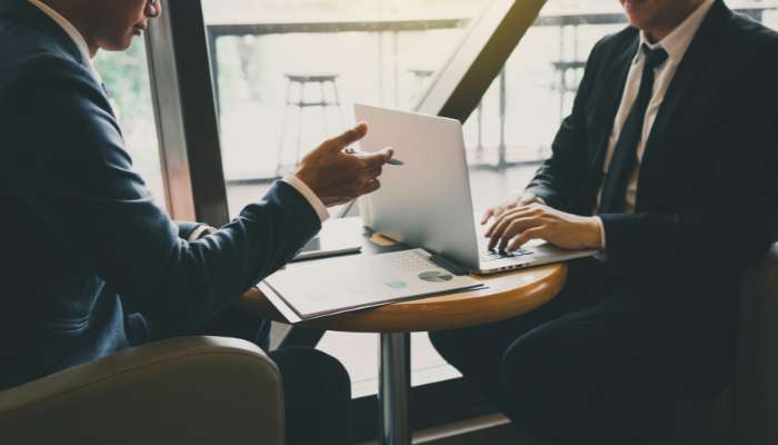 Two professionals in suits discuss at a small round table with an open laptop and documents nearby.