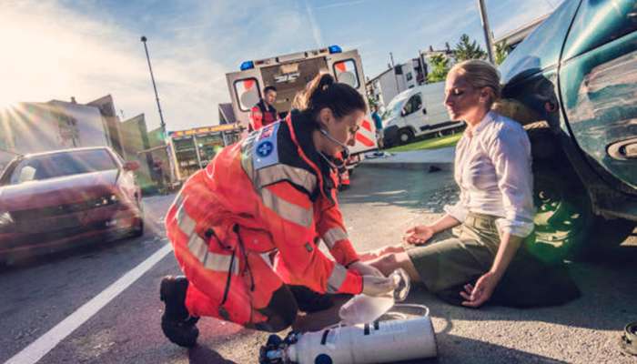 Paramedic kneels beside a woman on a road after a car crash, checking her vitals with an oxygen tank nearby.