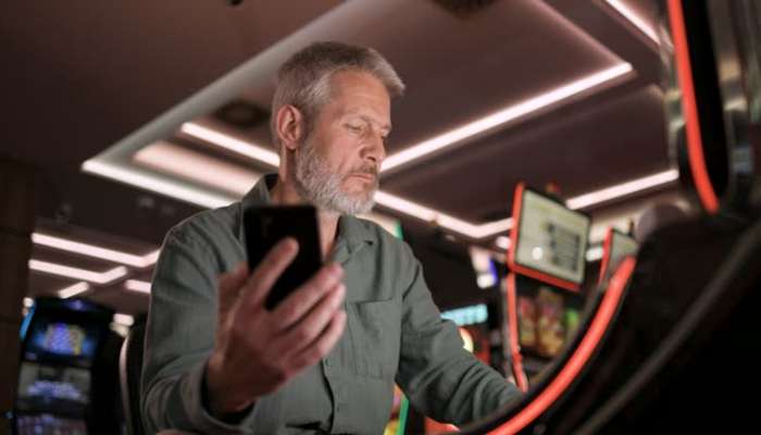Older man with gray beard looking at his phone while seated at a casino or arcade gaming machine area.