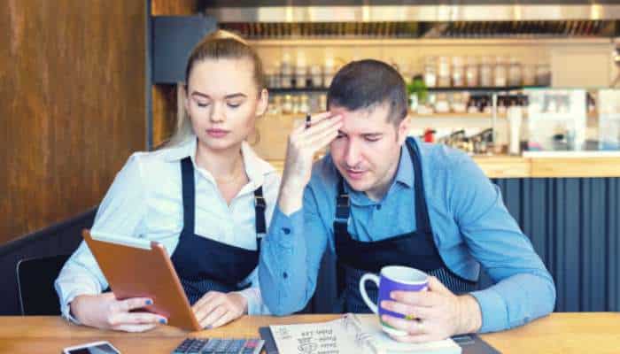 Why small business owners often underestimate their everyday operating costs 1 | tycoonstory media Two cafe workers in aprons study a clipboard and a calculator at the counter, looking focused.
