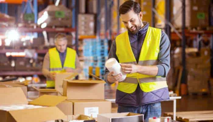 Two warehouse workers in yellow vests packing and inspecting boxes in a storage facility.