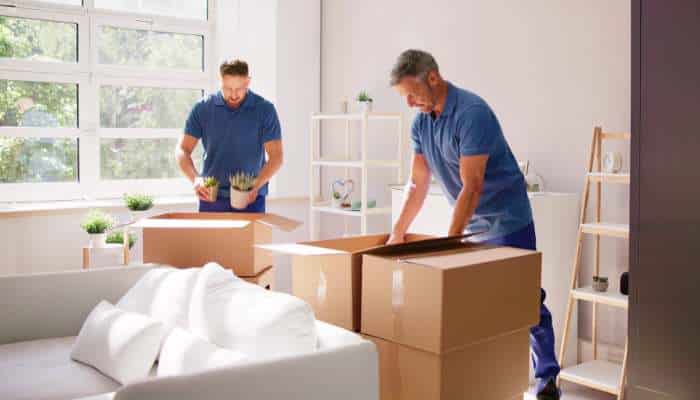 Why hiring experienced furniture movers makes a difference 1 | tycoonstory media Two movers in blue shirts packing boxes in a sunlit living room, with a sofa and shelves nearby.