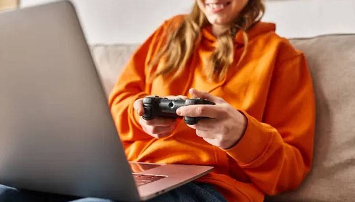 Woman in an orange hoodie using a controller and laptop for xbox cloud gaming, enjoying console-free gaming from the comfort of her home.