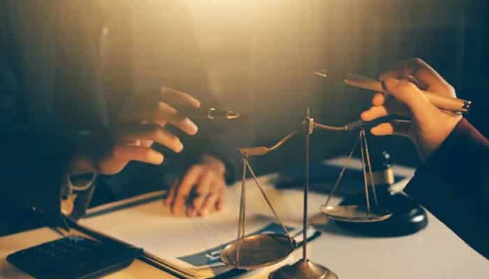 Two people discuss at a desk with a small balance scale and documents, suggesting a legal consultation.