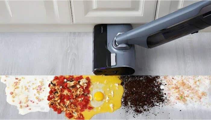 Vacuum cleaner head hovering over a line of spilled food on a light floor, including egg, cheese, sauce, and toppings by the cabinet.