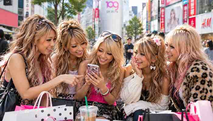 Gyaru culture beyond clothing and makeup - tycoonstory | tycoonstory media Gyaru meaning beyond fashion: five japanese gyaru-style women with voluminous hairstyles, dramatic makeup, and trendy outfits sharing a smartphone in tokyo's shibuya shopping district, highlighting friendship and youth culture.