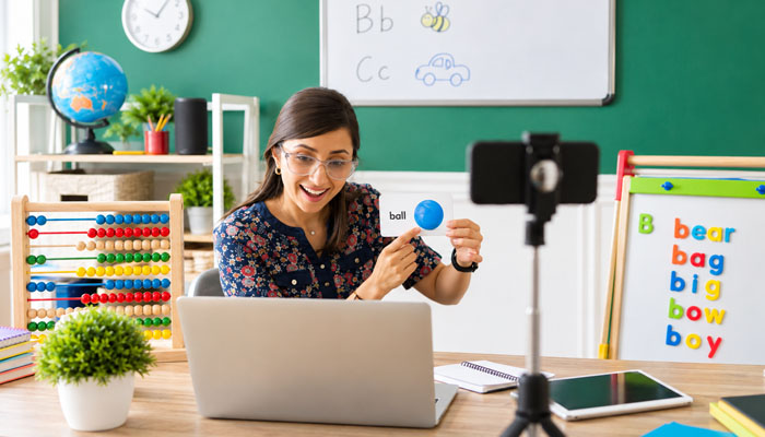 Teacher records a phonics lesson for kids, holding a blue ball and a card that says'ball' in a bright classroom setup with a laptop and teaching tools nearby.